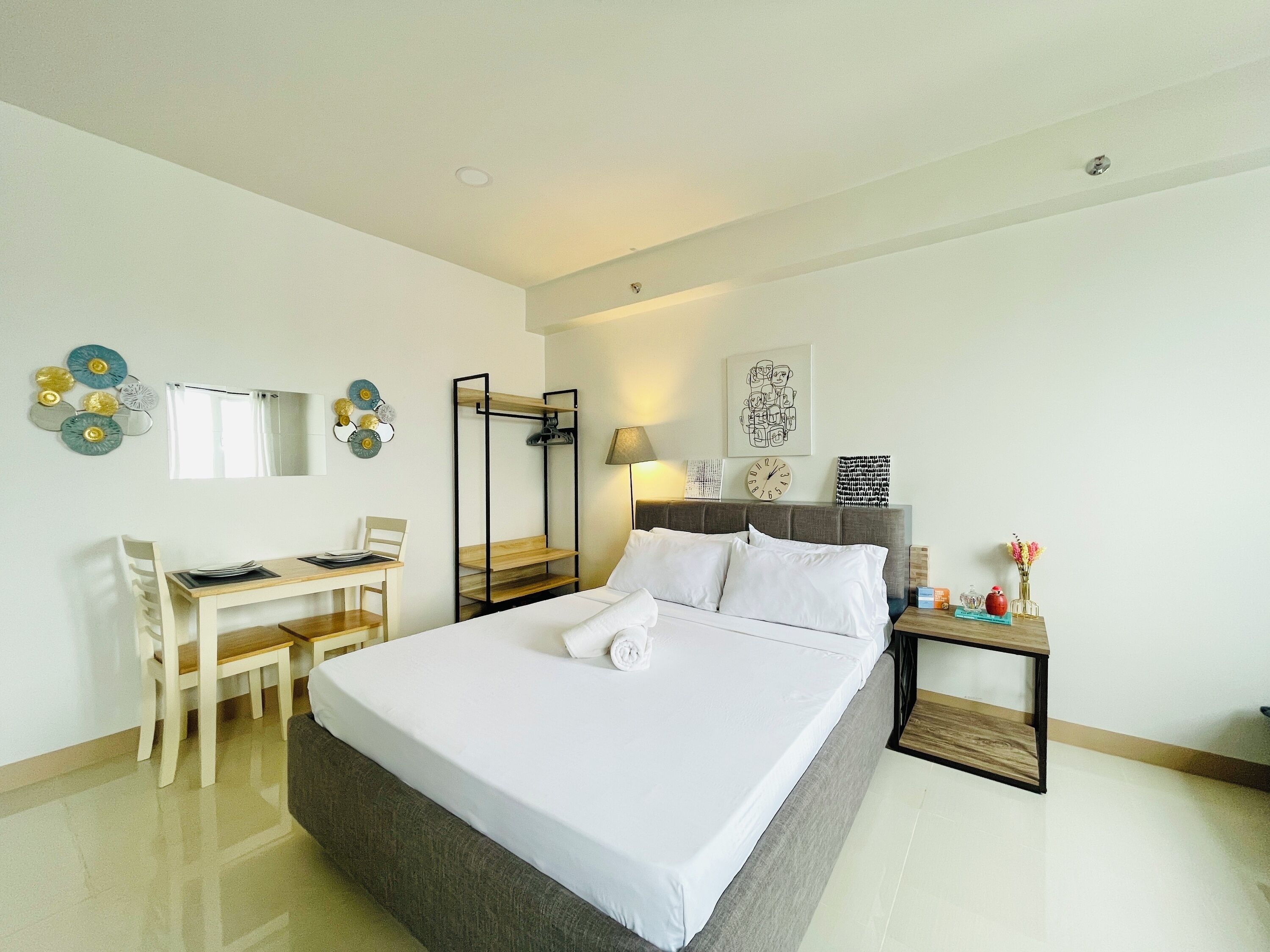 A hotel room with a bed, a desk, a chair, a small table, and a wall-mounted shelf.