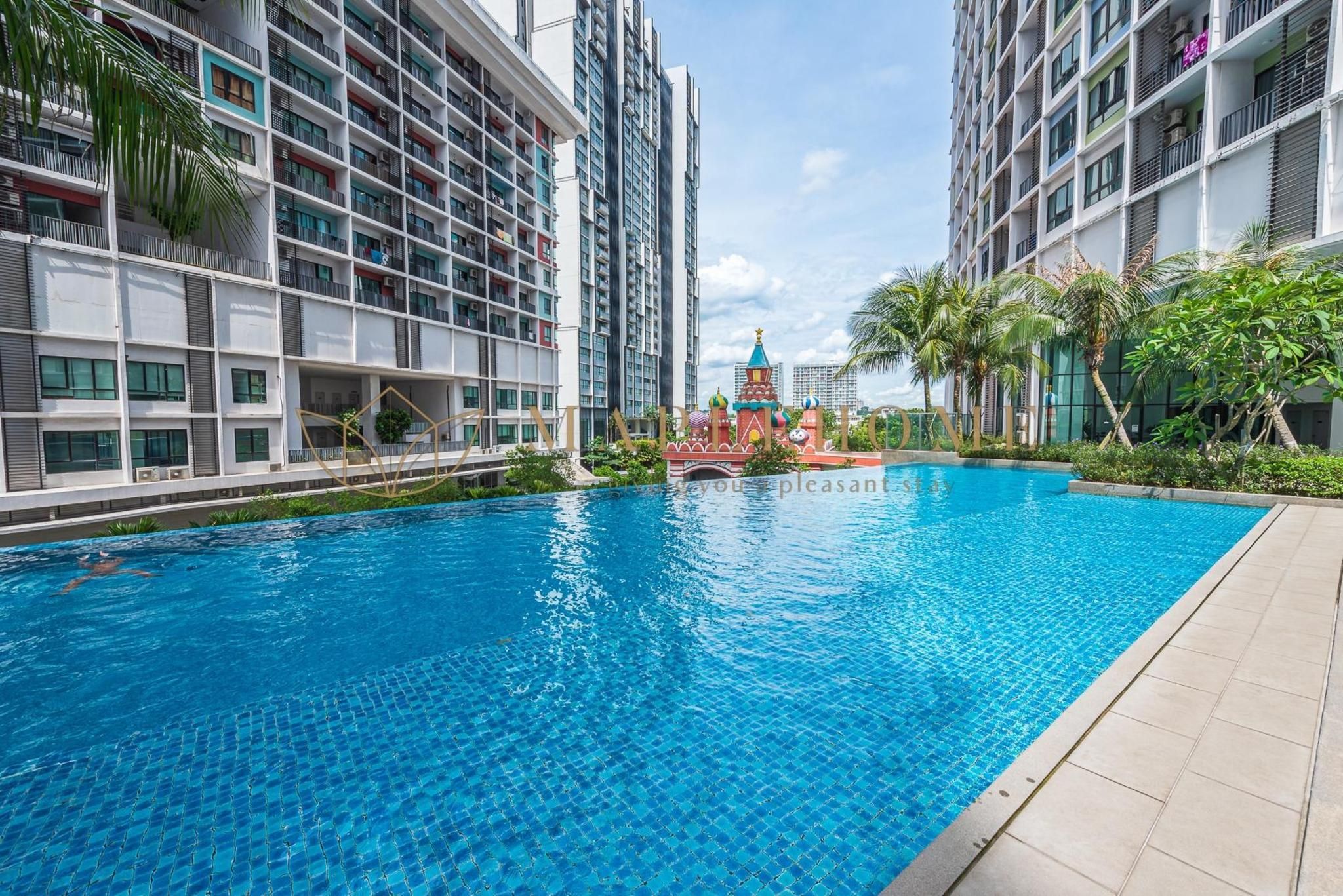 A swimming pool surrounded by modern high-rise buildings with balconies, a small bridge, and a decorative structure in the background.