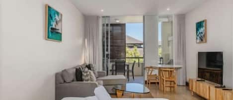 A modern hotel room with a bed, sofa, dining table, chairs, TV, and artwork.