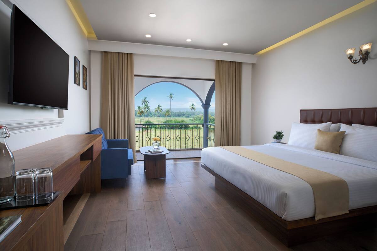 A hotel room with a large bed, a TV, a desk, and a balcony with a view of palm trees.
