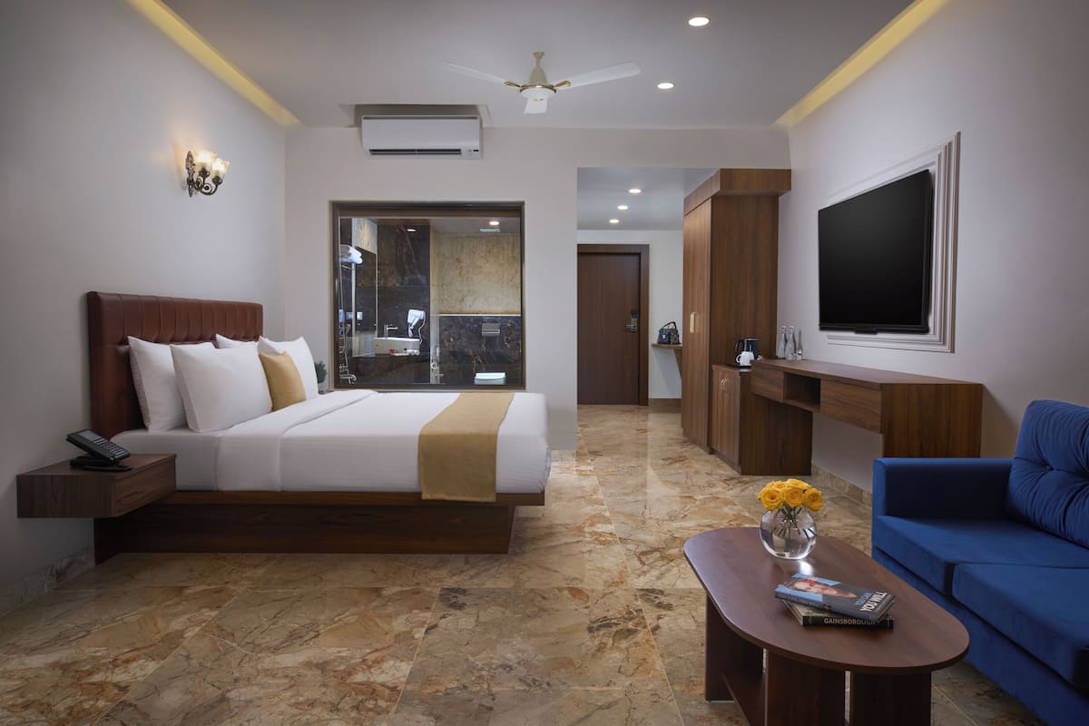 A modern hotel room with a bed, a sofa, a coffee table, a TV, and a bathroom visible through an open door.