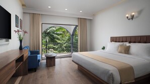 A modern hotel room with a large bed, a desk, a blue chair, and a balcony with a view of greenery. - Sahaswara Luxury Resort (Anjuna)