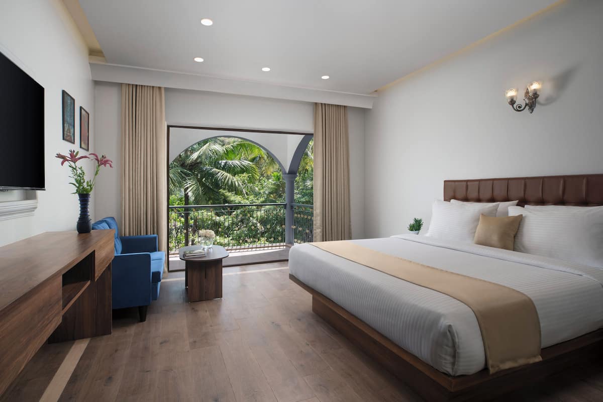 A modern hotel room with a large bed, a desk, a blue chair, and a balcony with a view of greenery.