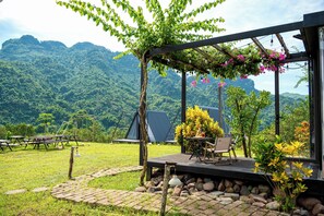A patio with a table and chairs, surrounded by a stone pathway and greenery, overlooking a mountainous landscape. - Ora Hill Farmstay & Glamping Hoa Binh (Thung Nai)