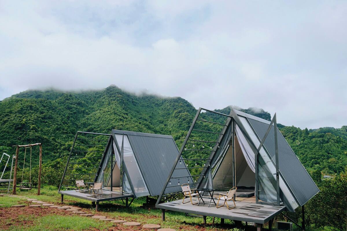 Dos tiendas modernas triangulares con paredes de vidrio, ubicadas en una zona cubierta de césped con una montaña boscosa al fondo.