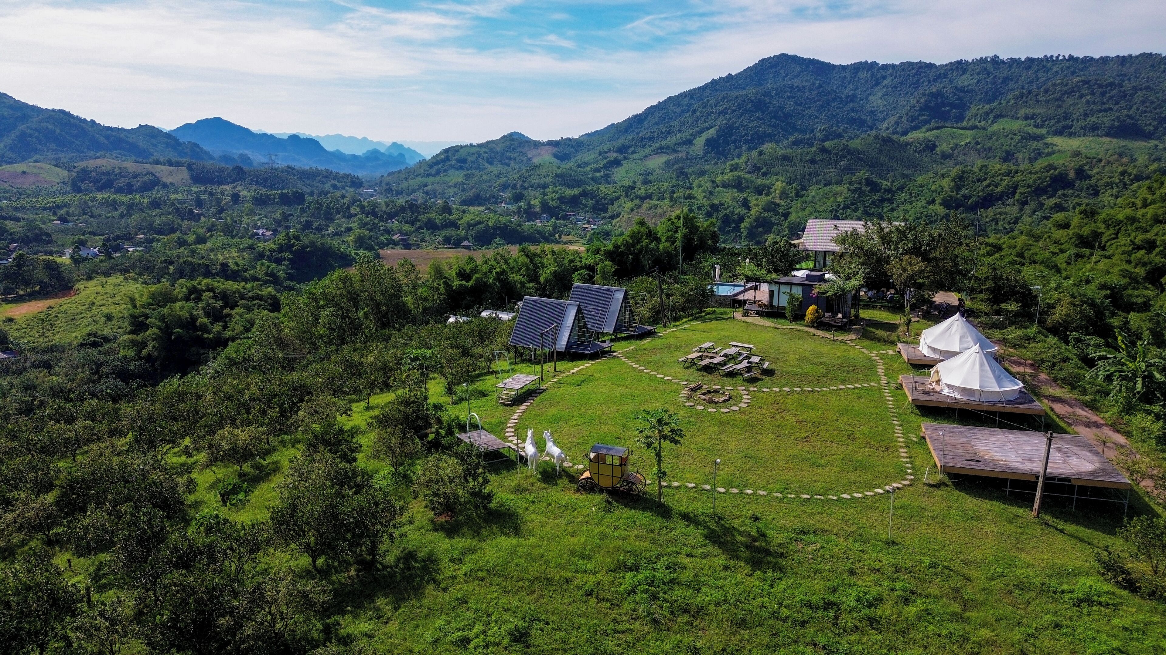 Un paysage rural avec un petit groupe de bâtiments, entouré d’une végétation luxuriante et dominé par des collines en arrière-plan.
