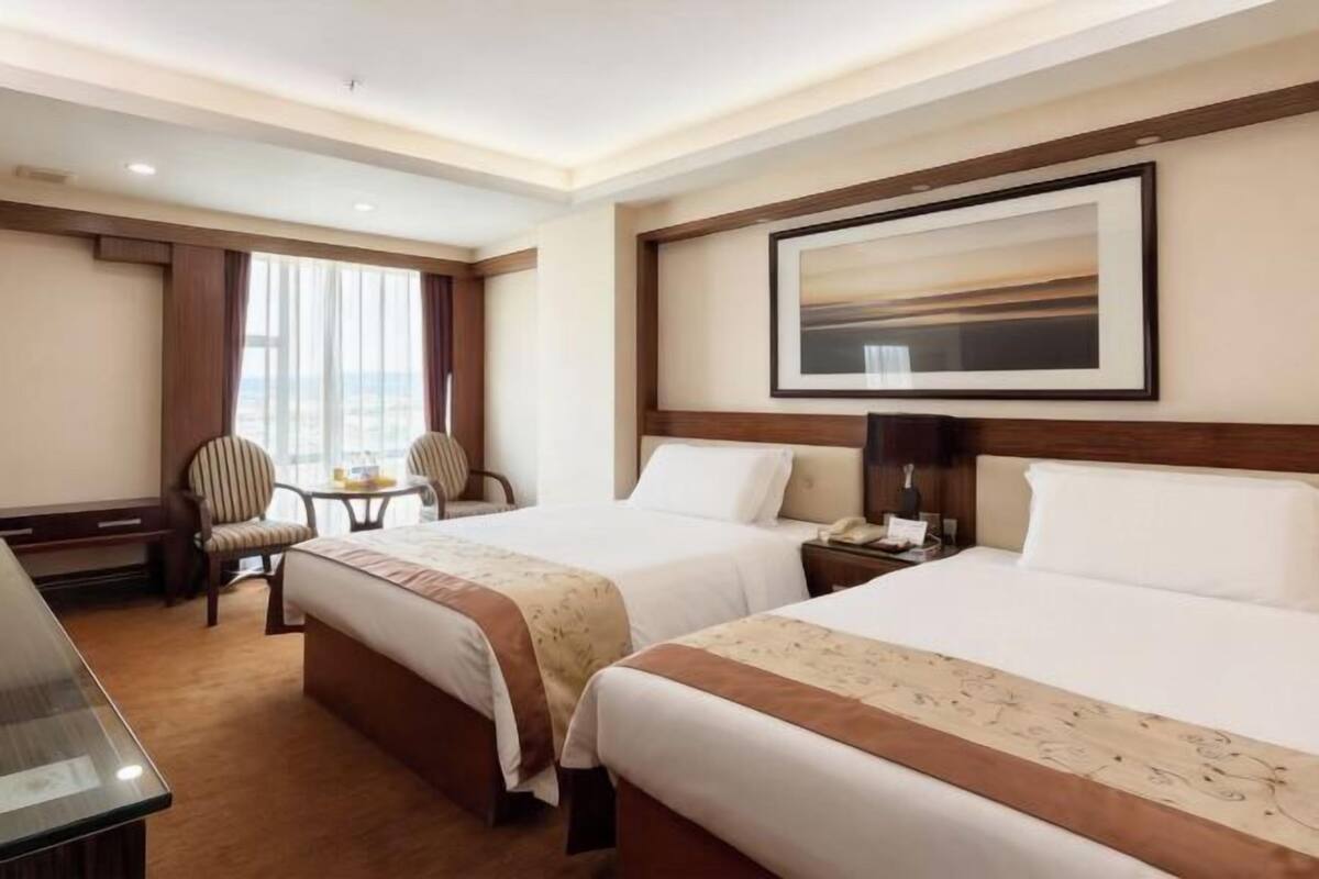 A hotel room with two beds, a desk, a chair, a window with curtains, and a framed picture on the wall.