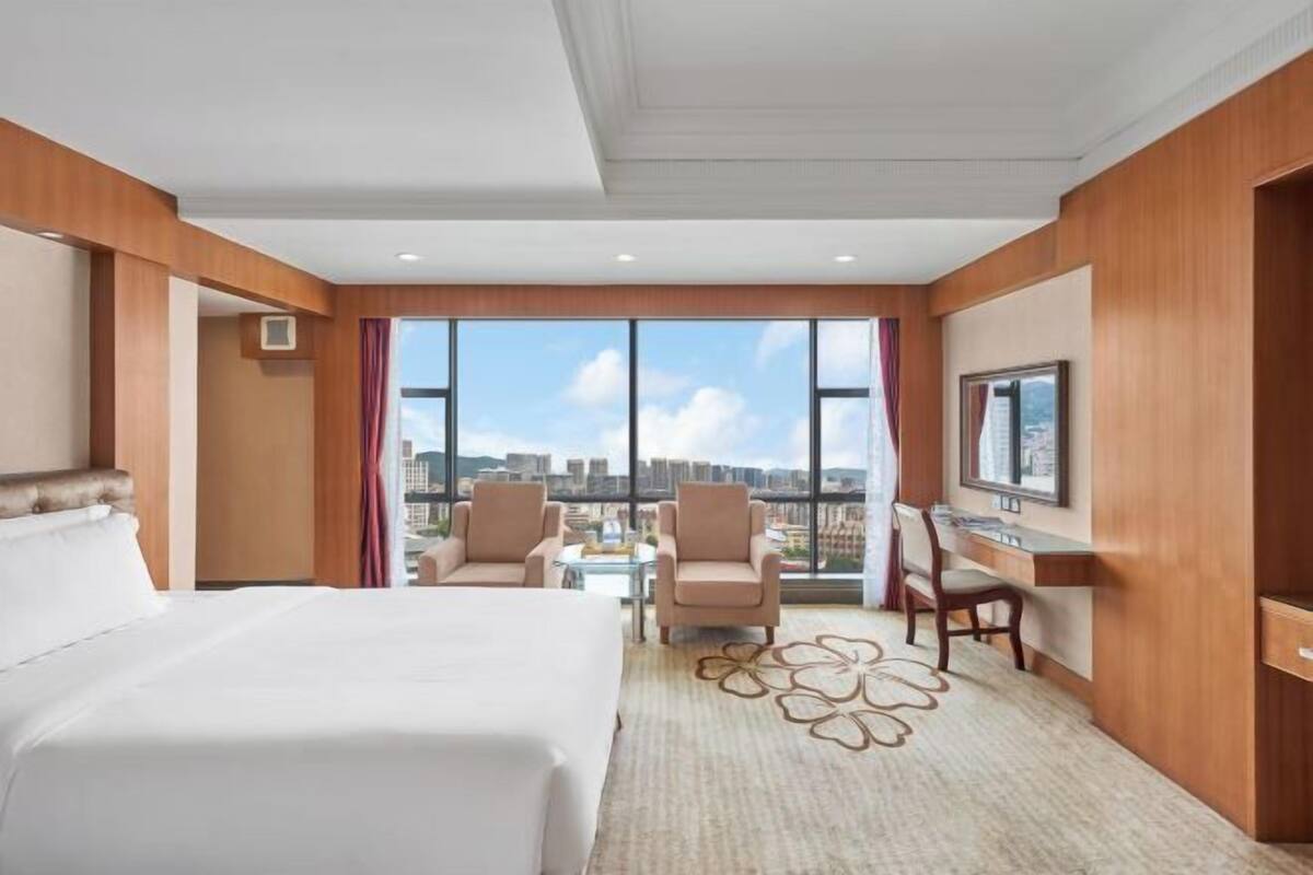 A hotel room with a large bed, two armchairs, a desk, and a TV. The room has a large window with a city view.