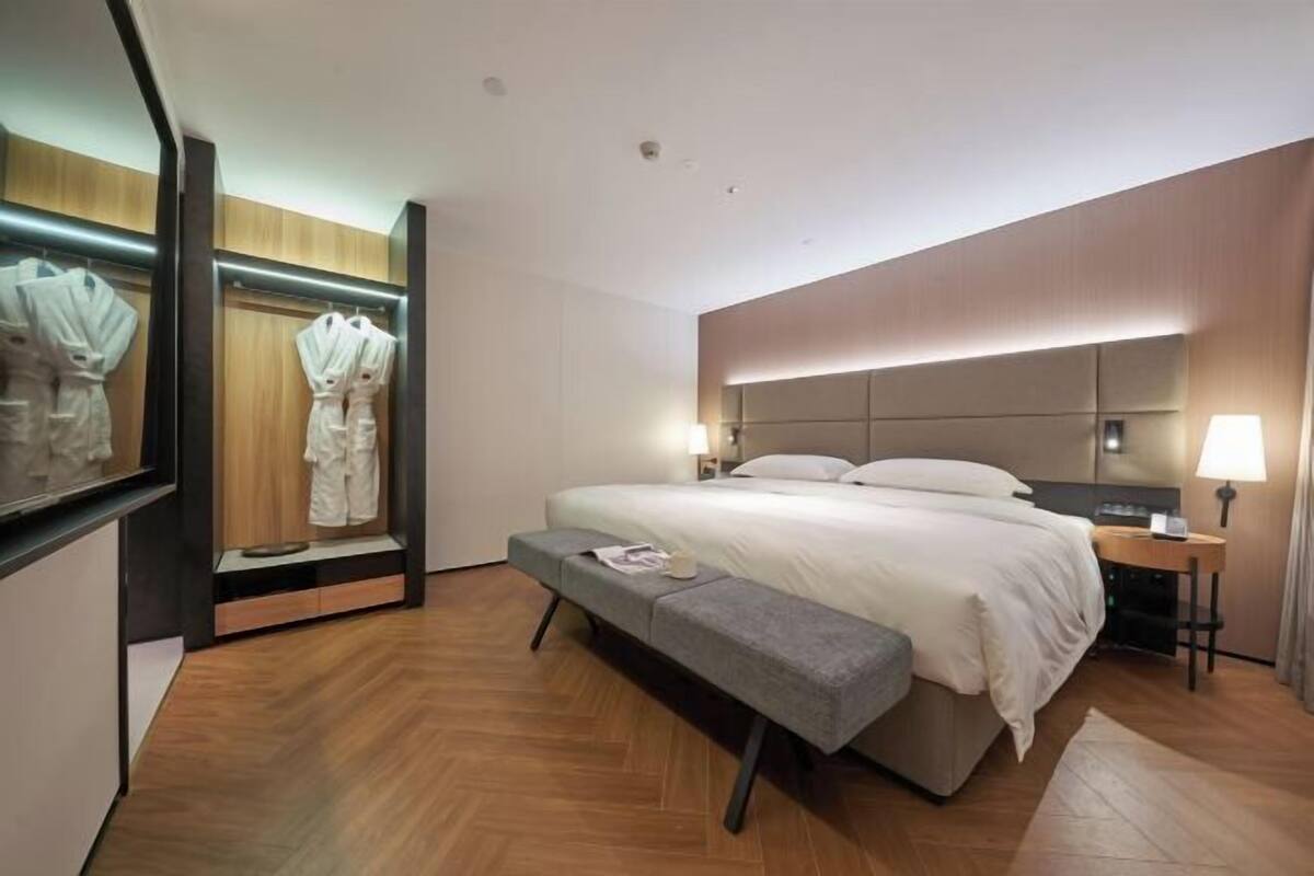 A modern hotel room with a large bed, bedside tables, a bench, a wardrobe, and a mirror.