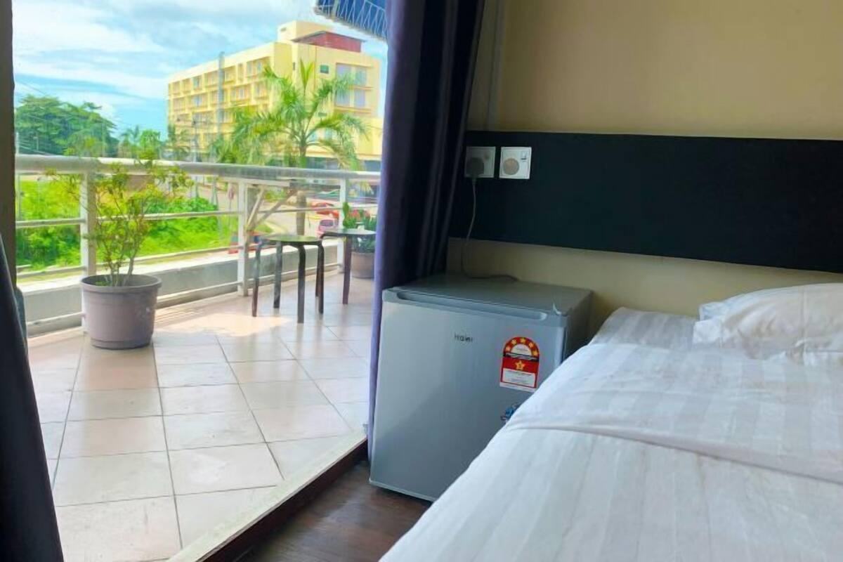 Habitación de hotel con cama, un refrigerador pequeño, un balcón con planta en maceta y vistas a edificios y árboles.