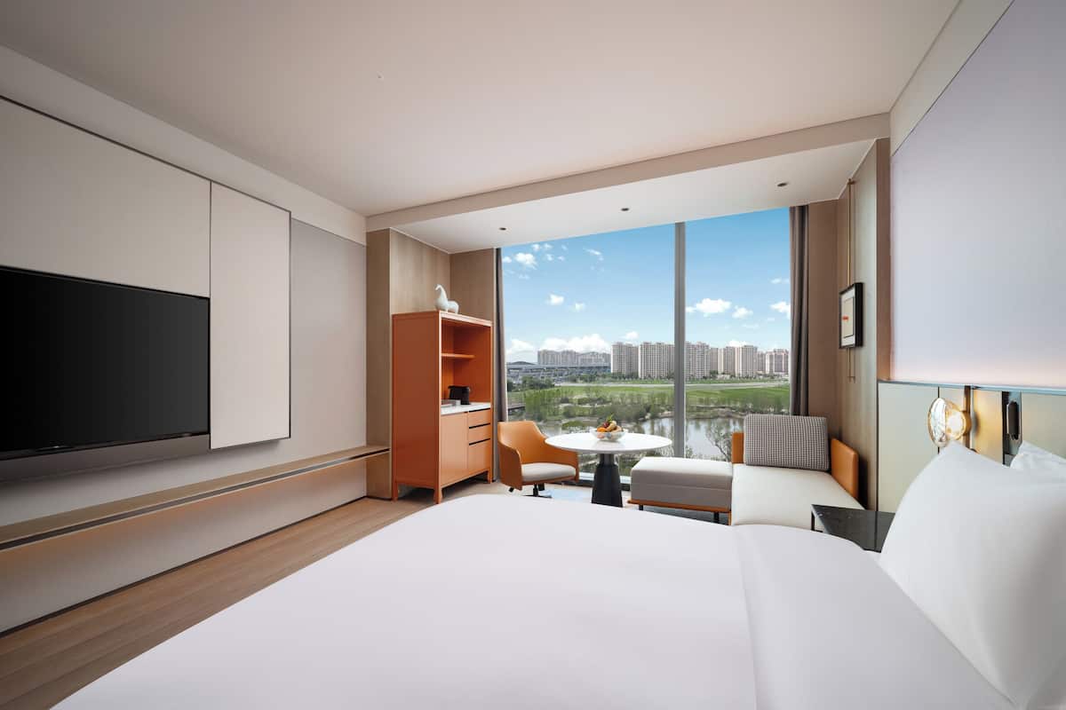 A modern hotel room with a large bed, a TV, a small table with a bowl of fruit, a sofa, and a view of the cityscape through large windows.