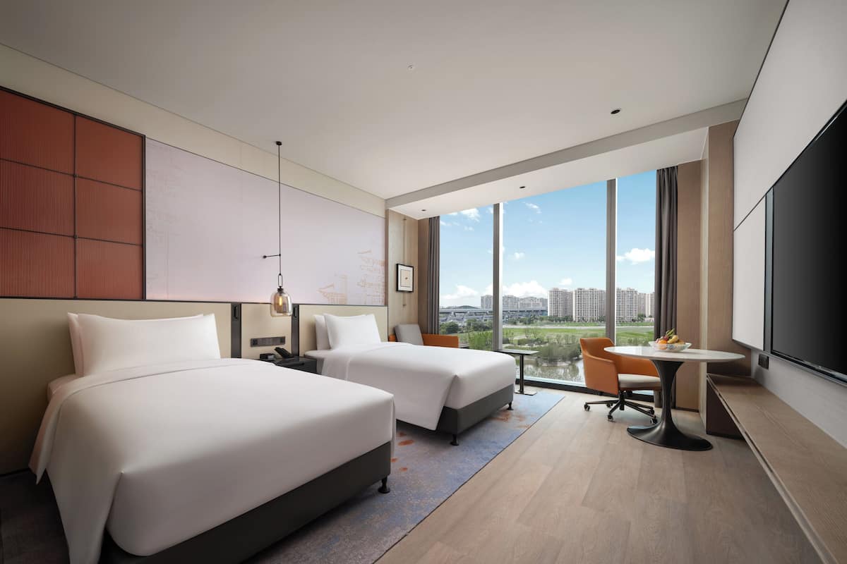 A modern hotel room with two beds, a desk, and a large window offering a city view.