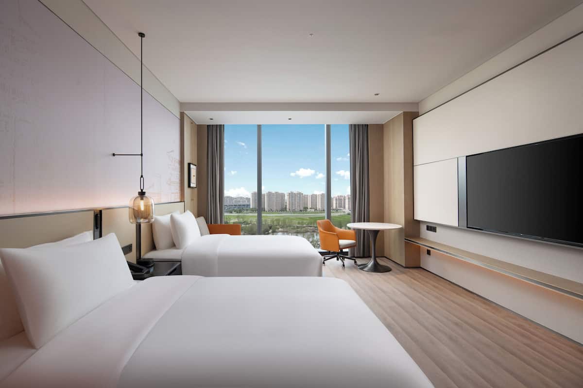 A modern hotel room with two beds, a TV, a desk, and a view of a cityscape.