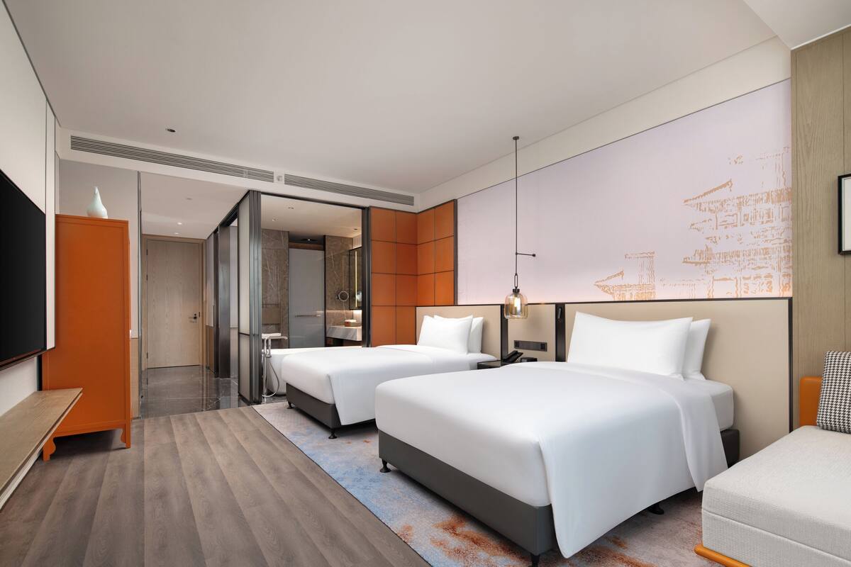 A modern hotel room with two beds, a flat-screen TV, and a large window.