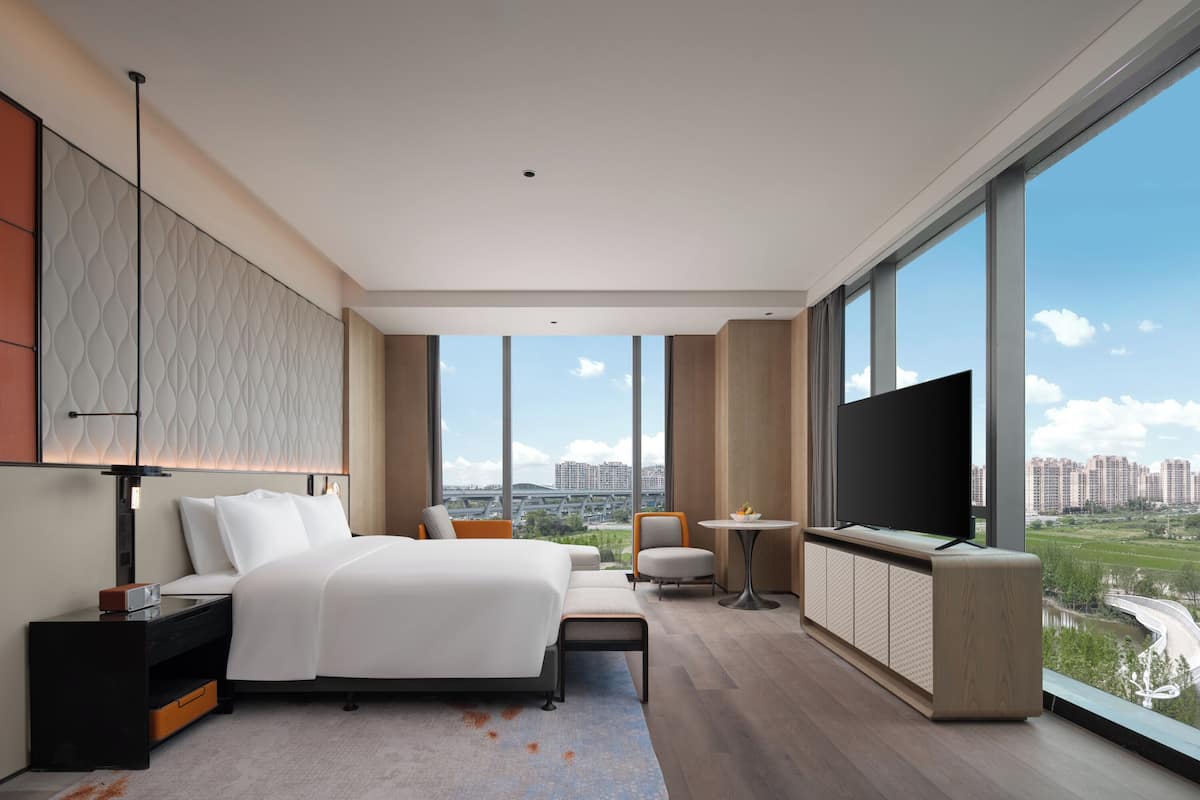 A modern hotel room with a large bed, a TV, a desk, and a view of the city.