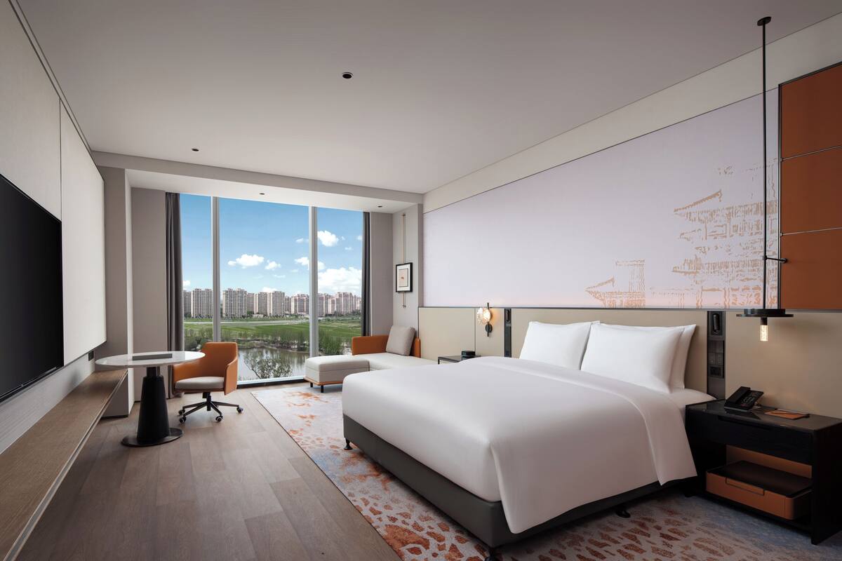 A modern hotel room with a large bed, a desk, a chair, and a view of the city through large windows.