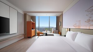 Una habitación de hotel moderna con una cama grande, televisor, mueble de madera, escritorio con silla y un ventanal con vistas a la ciudad.