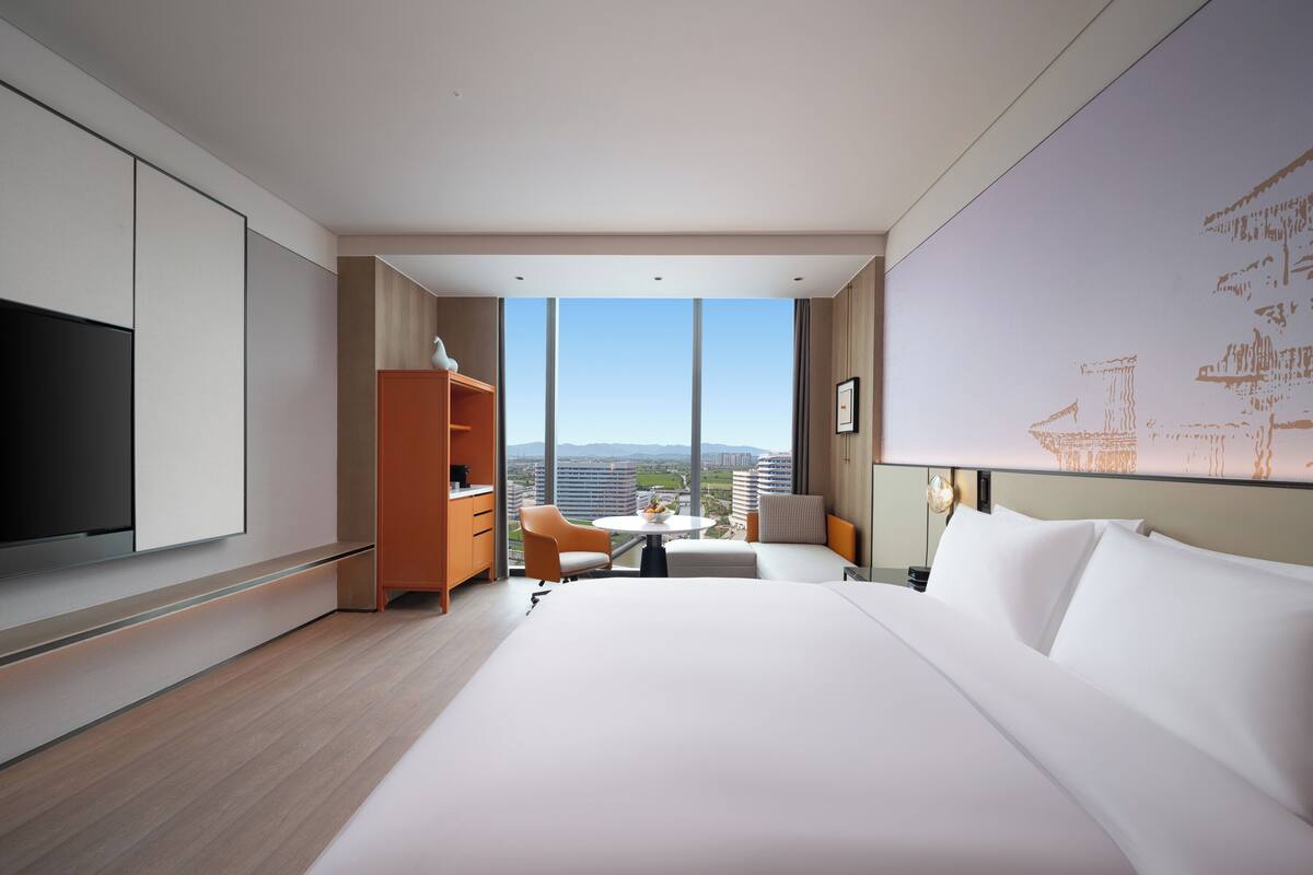 A modern hotel room with a large bed, a TV, a wooden cabinet, a desk with a chair, and a large window offering a city view.