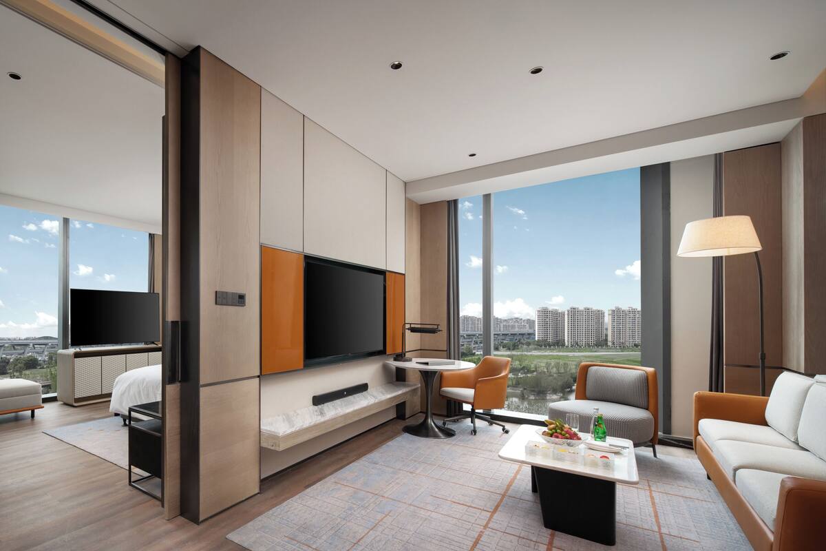 A modern hotel room with a large TV, comfortable seating, a dining area, and a view of the city.