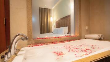 A hotel bathroom with a large bathtub filled with rose petals, a mirror, and a bed in the background.