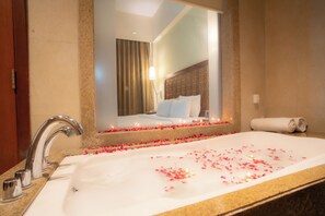 Un baño de hotel con una bañera grande llena de pétalos de rosa, un espejo y una cama al fondo.
