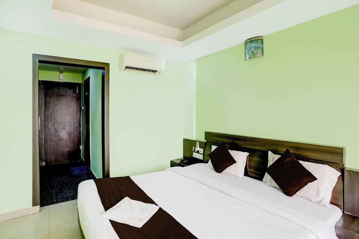 A hotel room with a bed, a nightstand, an air conditioner, and a door leading to another room.