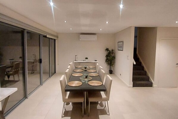 A dining area with a long table set for six, surrounded by chairs. There is a sliding glass door on one side, a staircase on the other, and a potted plant in the corner.