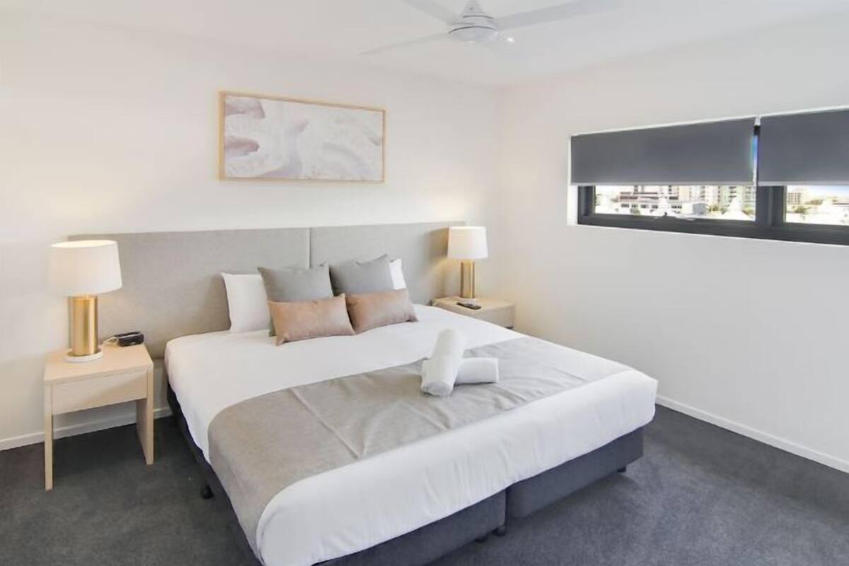 A hotel room with a bed, two bedside tables, a ceiling fan, a window with blinds, and a painting on the wall.