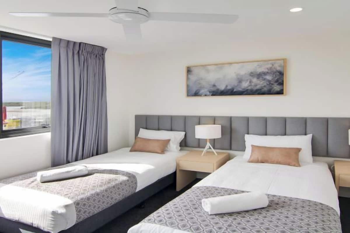 A hotel room with two beds, a ceiling fan, a window with curtains, a bedside table with a lamp, and a painting on the wall.