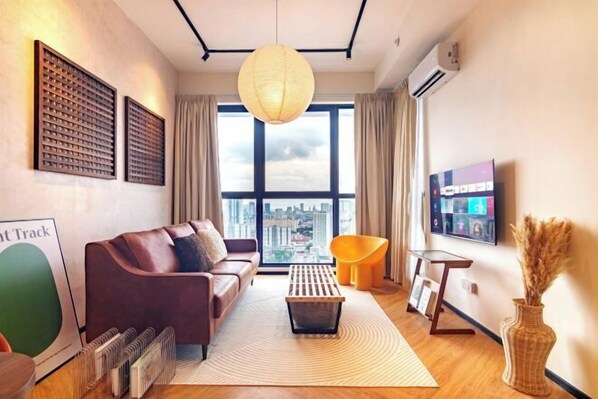 A modern living room with a sofa, a coffee table, a TV, and a large window with a city view. - Urban Suites by Stellar ALV (Jelutong)