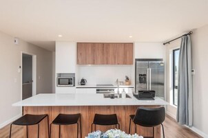 A modern kitchen with a central island, wooden cabinets, stainless steel appliances, and a large window with curtains.