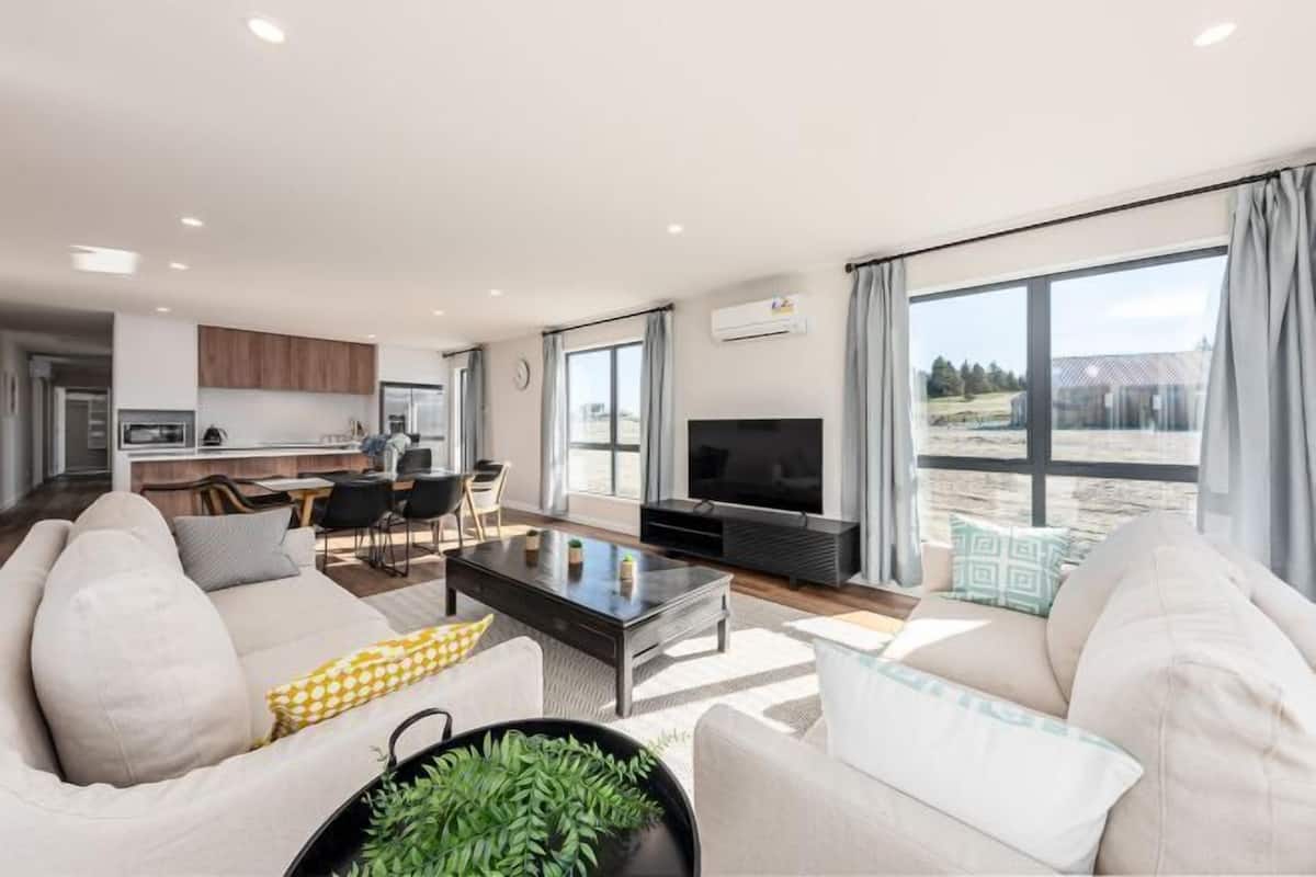A modern living room with a sofa, coffee table, and TV. There is a kitchen area in the background and a balcony with a view.