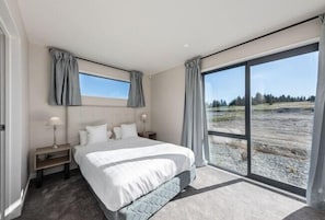 A modern bedroom with a large bed, a nightstand, a window with curtains, and a view of a natural landscape.