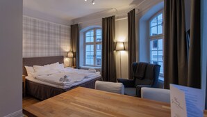A hotel room with a bed, a wooden table, two chairs, a desk, a lamp, and a window with curtains.