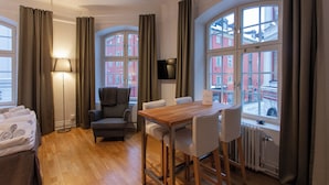 A hotel room with a wooden floor, a bed with neatly folded towels, a grey armchair, a wooden table with chairs, a floor lamp, and large windows with curtains.