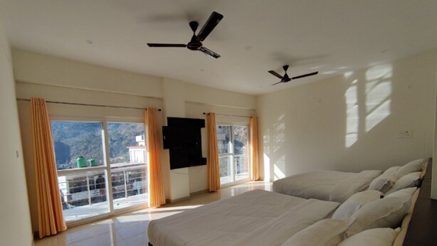 A bedroom with two beds, ceiling fans, a TV, and a balcony with curtains. - Hotel Nirvanam (Chamoli)