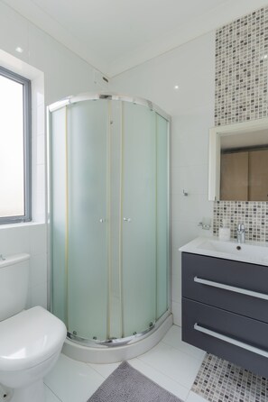 Un baño moderno con ducha envuelta en vidrio, inodoro blanco, tocador oscuro con cajones y salpicadero de azulejos.