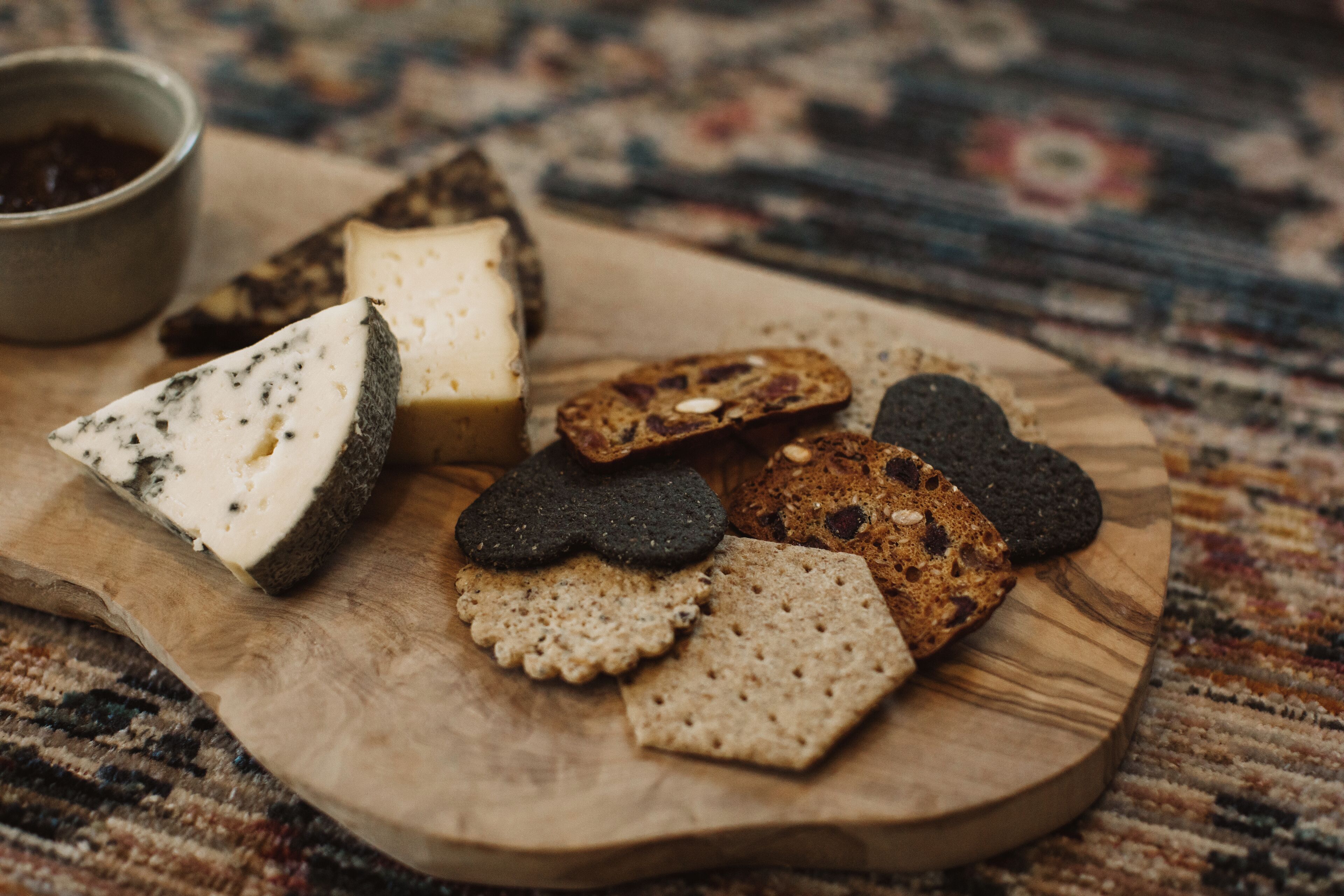 Una tabla de madera con varios quesos, galletas y un pequeño cuenco de mermelada.