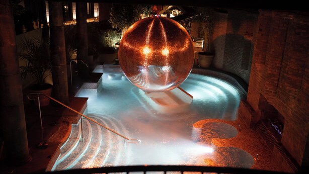 A spherical water feature in a pool with a brick wall and potted plants in the background.