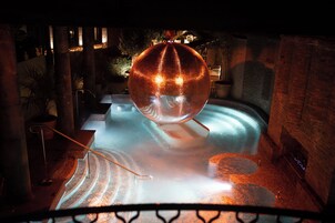 A spherical water feature in a pool with a brick wall and potted plants in the background.
