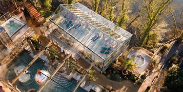 A glasshouse structure with a pool and outdoor seating area surrounded by trees.