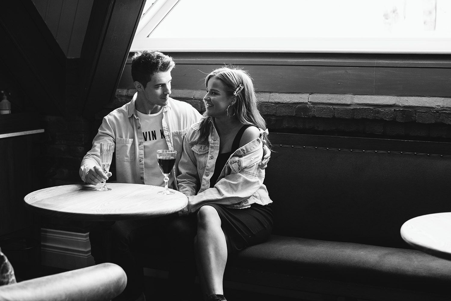 Una pareja sentada en una mesa redonda en una habitación con poca luz, pared de ladrillo y una ventana.