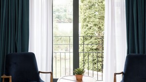 A hotel room with a bed, two chairs, a small table with a plant, and a view of a balcony with greenery.
