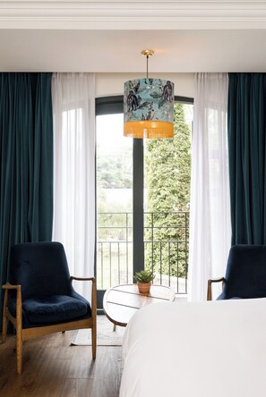A hotel room with a bed, two chairs, a small table with a plant, and a view of a balcony with greenery.