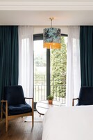 A hotel room with a bed, two chairs, a small table with a plant, and a view of a balcony with greenery.