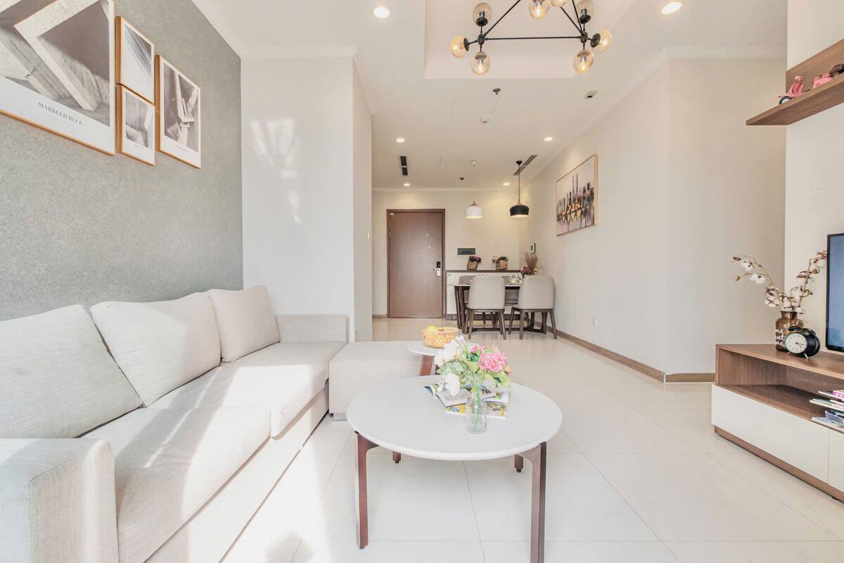 A modern living room with a white sofa, a round coffee table with flowers, and a dining area in the background.