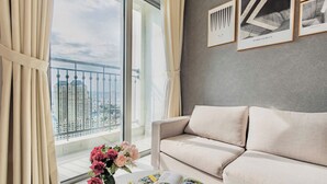 A modern living room with a beige sofa, a coffee table with a magazine, a vase with flowers, and a large window with a view of high-rise buildings.
