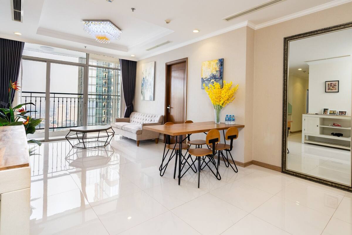 A modern hotel room with a dining area, a sofa, a large mirror, and a balcony with a view of the city.