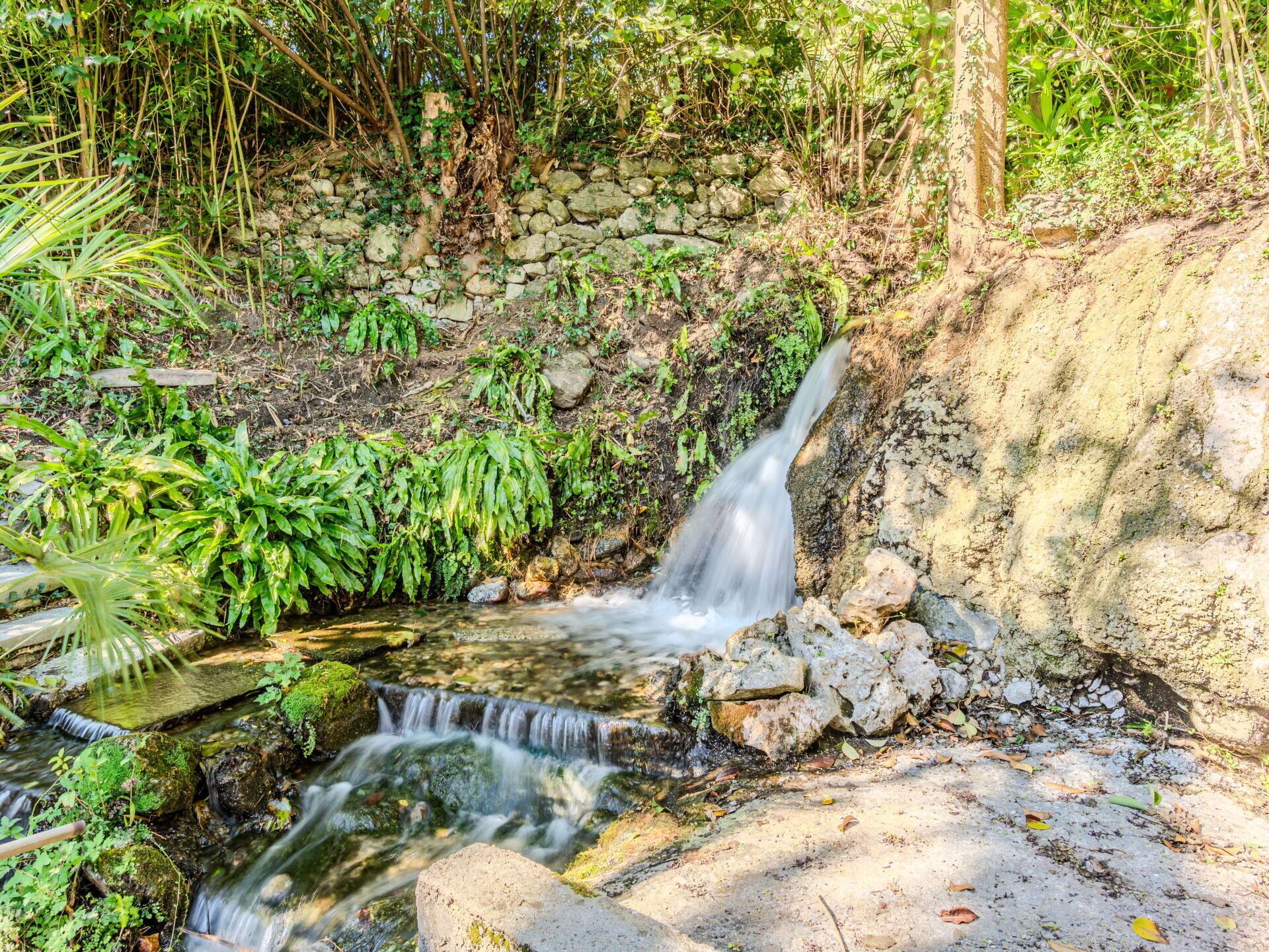 Ein kleiner Wasserfall in einer natürlichen Umgebung mit umgebendem Grün und einer Steinmauer.