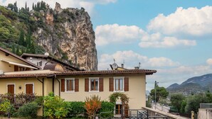 A building with a stone staircase leading to a door, a garden with various plants, and a cliff in the background. - Beautiful apartment for 6 guests with TV and patio (Arco)